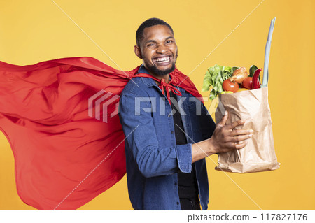 African american adult smiling and acting like a superhero with a bag of freshly harvested vegetables, promoting sustainable lifestyle. Young man recommending vegan nutrition in studio. 117827176
