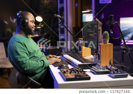 African american producer mixing sounds and tunes on his audio files, listening to the final product on headset. Audio technician works on pc with DAW software interface and stereo equipment. African american producer mixing sounds and tunes on his audio files, listening to the final product on headset. Audio technician works on pc with DAW software interface and stereo equipment. 117827188