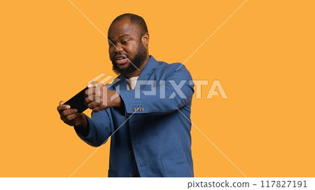 African american man playing videogames on cellphone in landscape mode, enjoying leisure time. Gamer enjoying game on mobile phone, having fun, studio background, camera A African american man playing videogames on cellphone in landscape mode, enjoying leisure time. Gamer enjoying game on mobile phone, having fun, studio background, camera A 117827191