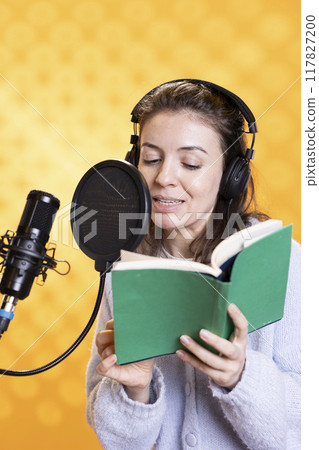Smiling narrator wearing headphones reading aloud from book into microphone against yellow background. Cheerful person recording audiobook, creating engaging media content for listeners Smiling narrator wearing headphones reading aloud from book into microphone against yellow background. Cheerful person recording audiobook, creating engaging media content for listeners 117827200