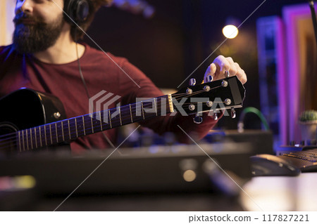Sound engineer artist tuning his acoustic guitar strings to play and record new tracks, working with electronic mixing and mastering gear in home studio. Young musician creating cool tunes. 117827221