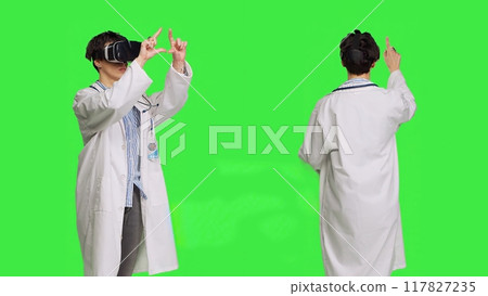 Woman medic works with 3d virtual reality headset in studio, standing against greenscreen backdrop. General practitioner uses vr glasses with interactive vision tool, futuristic exam. Camera B. 117827235