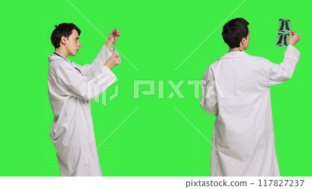 General practitioner examining radiography scan against greenscreen backdrop, looking at x ray results to find diagnosis. Physician in white coat uses radiology science and research. Camera B. 117827237
