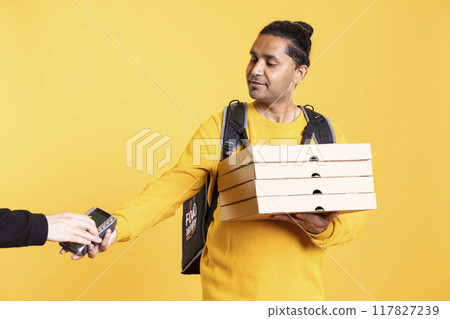 Smiling delivery worker offering takeaway order to customer paying with debit card. Client buying meal from courier, swiping card on POS device, isolated over studio background 117827239