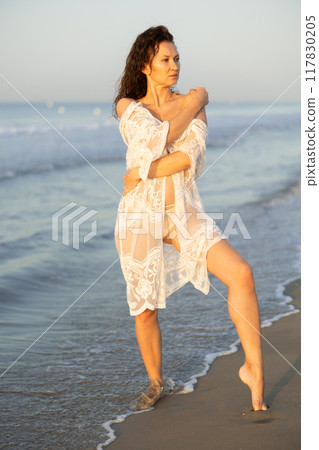 Sexy woman in white lingerie and mesh cape walking along the seashore in surf at sunset 117830205