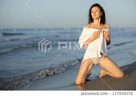 Topless woman in white shirt kneeling on coast 117830258