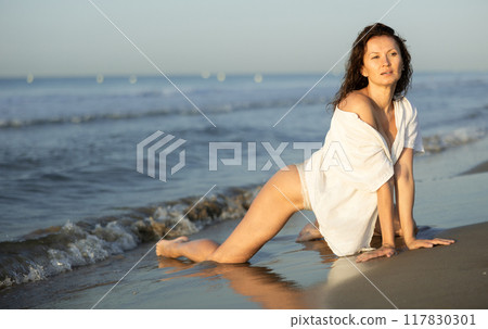 Portrait of female posing in white lingerie and mesh cape on knees on beach near sea at sunset Portrait of female posing in white lingerie and mesh cape on knees on beach near sea at sunset 117830301