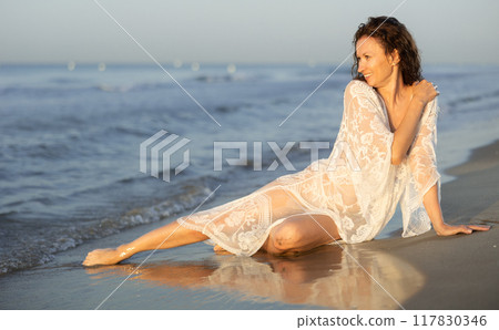 Woman in white lingerie and mesh cape sitting in rays of setting sun on seashore Woman in white lingerie and mesh cape sitting in rays of setting sun on seashore 117830346