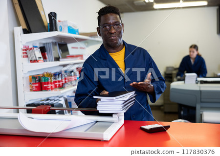 Portrait of friendly male employee of printing house with stack of notebooks in her hands Portrait of friendly male employee of printing house with stack of notebooks in her hands 117830376