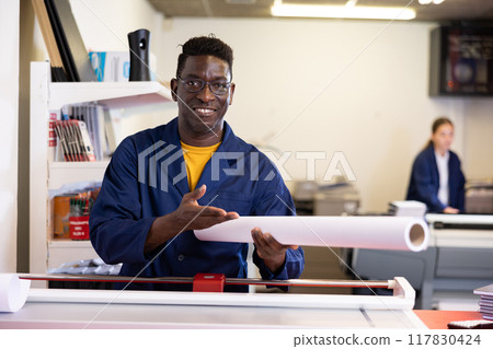 Middle-aged African American male specialist in uniform holding whatman in a roll in the middle of print shop 117830424