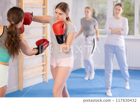 Two teenage girls in sportswear are engaged in boxing sparring in the gym under guidance of trainer in gym Two teenage girls in sportswear are engaged in boxing sparring in the gym under guidance of trainer in gym 117830432