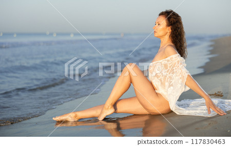 Adult woman in white peignoir sitting on coast Adult woman in white peignoir sitting on coast 117830463