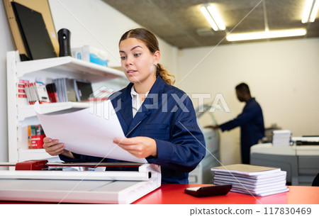 Serious young woman in a blue uniform using paper cutter on the table with planners and calculator in the printer house 117830469