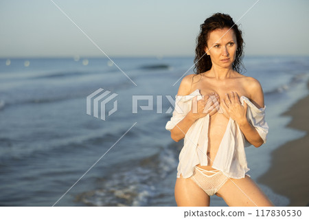 Sexy woman in white lingerie and mesh cape walking along the seashore in surf at sunset Sexy woman in white lingerie and mesh cape walking along the seashore in surf at sunset 117830530