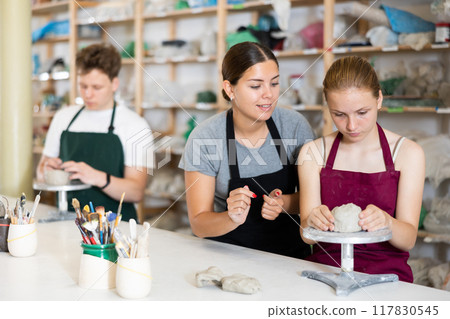 Young woman teacher helps teenagers boy and girl to sculpt ceramic product Young woman teacher helps teenagers boy and girl to sculpt ceramic product 117830545