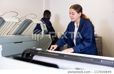 Focused young woman in uniform setting up the plotter before starting work in the printing house Focused young woman in uniform setting up the plotter before starting work in the printing house 117830649
