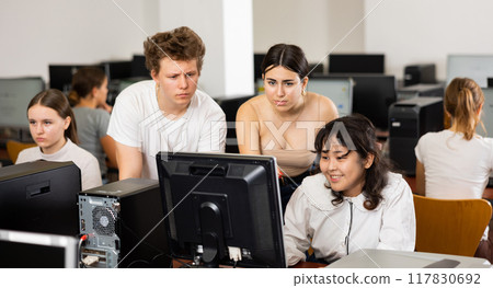 Teenage boy sitting at table and using computer during computer science lesson. Teacher and classmates standing and looking at monitor 117830692