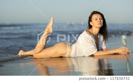 Topless woman in white cape with glass of wine on seashore Topless woman in white cape with glass of wine on seashore 117830731