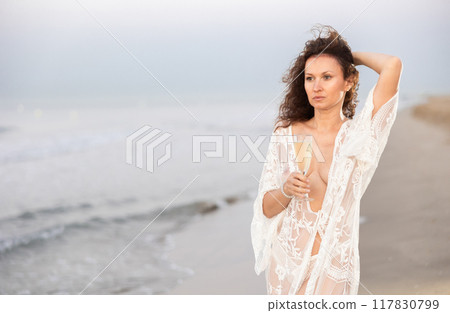 Portrait of woman in white mesh cape with glass of champagne posing on seashore 117830799
