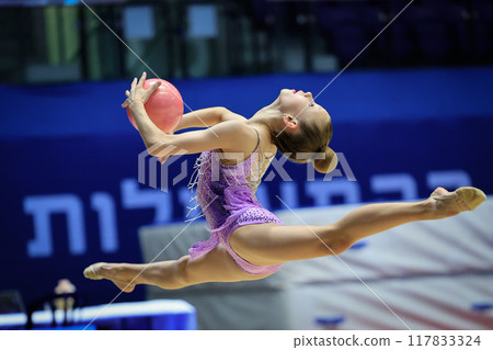 A young gymnast is showcasing a wonderful jump routine with a ball during a competitive event A young gymnast is showcasing a wonderful jump routine with a ball during a competitive event 117833324