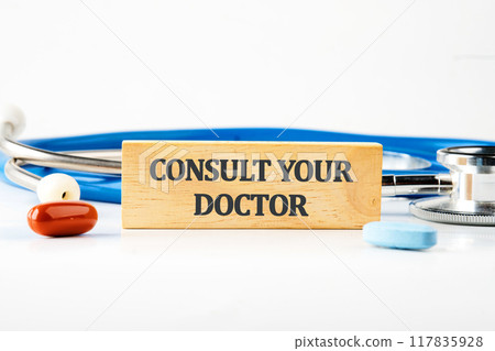 Medicine concept. Text CONSULT YOUR DOCTOR written on a wooden block with a stethoscope lying next to it, pills, vitamins on a white background 117835928