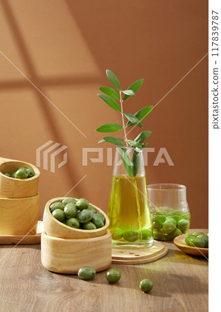 Fresh green olives are placed in wooden bowls, decorated with a vase of olive oil and green leaves on a wooden table and brown background. Front view. Olive oil is protective against heart disease 117839787