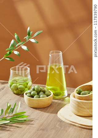 Minimal concept for advertising product with ingredient from olive. On wooden table and brown background, fresh olives, leaves and flask of olive oil decorated Minimal concept for advertising product with ingredient from olive. On wooden table and brown background, fresh olives, leaves and flask of olive oil decorated 117839790