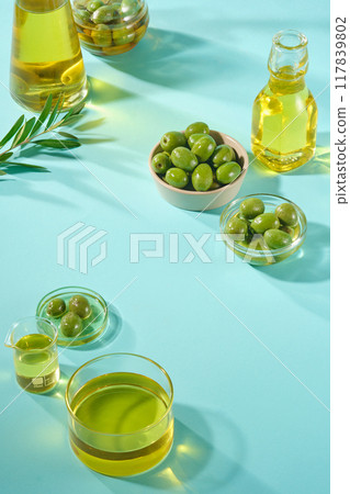 Minimalist background for cosmetic presentation with olive ingredient. Glass bottles of olive oil, fresh olives and leave decorated on blue background. Front view. Concept for natural extract. Minimalist background for cosmetic presentation with olive ingredient. Glass bottles of olive oil, fresh olives and leave decorated on blue background. Front view. Concept for natural extract. 117839802