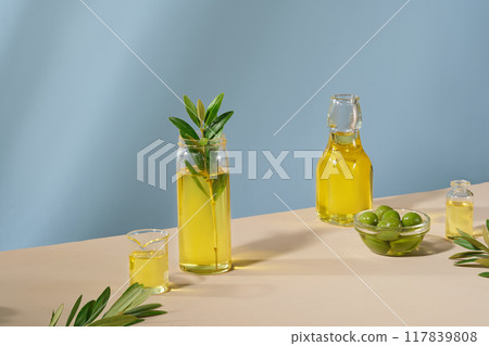 Minimal scene with blank space for show your product. Glass bottles of olive oil, fresh olives and leaves displayed on blue background. Minimal scene with blank space for show your product. Glass bottles of olive oil, fresh olives and leaves displayed on blue background. 117839808