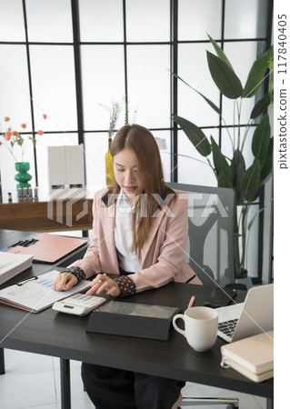 An Asian businesswoman writes on a digital tablet at her desk in a modern office. She is focused on her work, with documents and office supplies 117840405