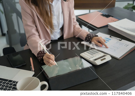 An Asian businesswoman writes on a digital tablet at her desk in a modern office. She is focused on her work, with documents and office supplies An Asian businesswoman writes on a digital tablet at her desk in a modern office. She is focused on her work, with documents and office supplies 117840406