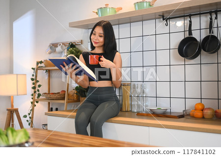 Pleased millennial woman in activewear reading book in minimal kitchen interior Pleased millennial woman in activewear reading book in minimal kitchen interior 117841102