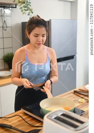 Beautiful young woman in sports wear frying eggs in a cooking pan in domestic kitchen 117841119