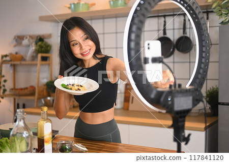 Young slim sportswoman making a healthy and recording video lesson on smart phone Young slim sportswoman making a healthy and recording video lesson on smart phone 117841120