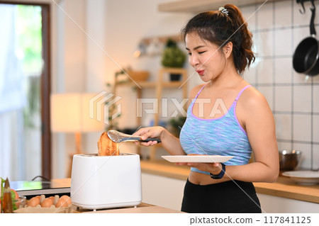 Shot pretty young woman in sports clothing toasting bread with toaster in the kitchen 117841125