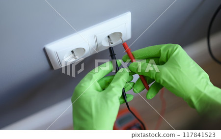 Hands in green gloves plug voltmeter into an outlet in wall Hands in green gloves plug voltmeter into an outlet in wall 117841523