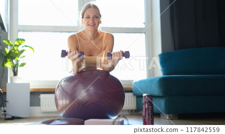 Woman doing exercises with dumbbells on a burgundy ball Woman doing exercises with dumbbells on a burgundy ball 117842559