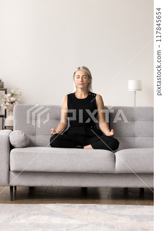 Serene woman doing meditation seated in lotus position on sofa 117845654