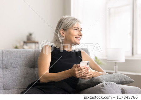 Pensive mature 50s woman sits on couch with smartphone 117845760