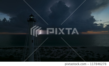 White tall lighthouse on sea shore with blinking light at stormy night for commercial vessels navigation. Thunderstorm with lightnings over ocean water posing danger for ships White tall lighthouse on sea shore with blinking light at stormy night for commercial vessels navigation. Thunderstorm with lightnings over ocean water posing danger for ships 117846278