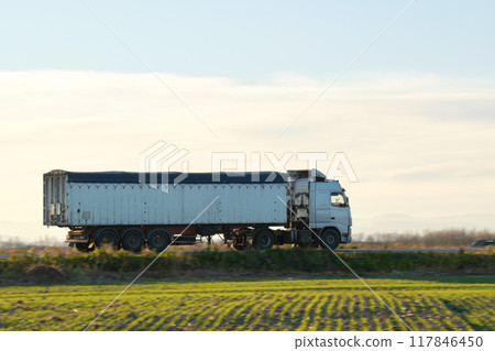 Semi-truck with tipping cargo trailer transporting sand from quarry driving on highway hauling goods in evening. Delivery transportation and logistics concept Semi-truck with tipping cargo trailer transporting sand from quarry driving on highway hauling goods in evening. Delivery transportation and logistics concept 117846450