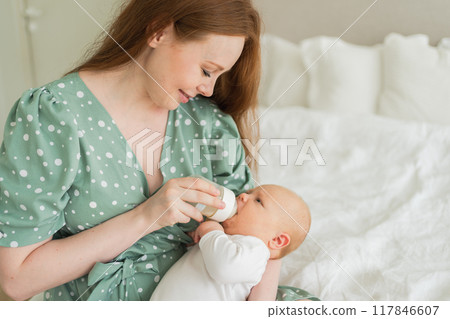Happy family at home. Mother holding little baby toddler child feeding milk from bottle. Newborn Infant baby girl sucking eating drinking milk. Mom of breast feeding baby. Motherhood happy child 117846607