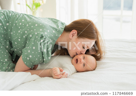 Happy family at home. Mother playing with little toddler child on bed. Mom baby relax playing having fun together. Mother looks at baby with love care. Mom of breast feeding baby rest in living room 117846669