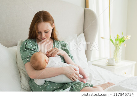 Happy family at home. Mother breastfeeding baby for health nutrition wellness. Mom nursing feeding her newborn child milk at home. Woman infant girl in white bedroom Mother hugs baby with love care 117846734
