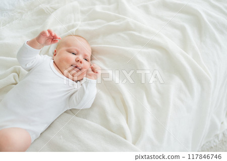 Happy baby. Cute little newborn girl with smiling face lying on bed in bedroom. Infant baby resting playing lying down on white blanket at home. Motherhood happy child childcare concept 117846746