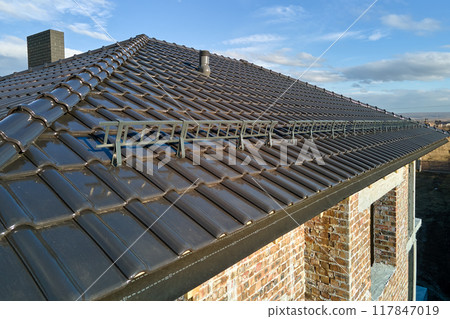 Closeup snow guard for safety in winter on house roof top covered with ceramic shingles. Tiled covering of building 117847019