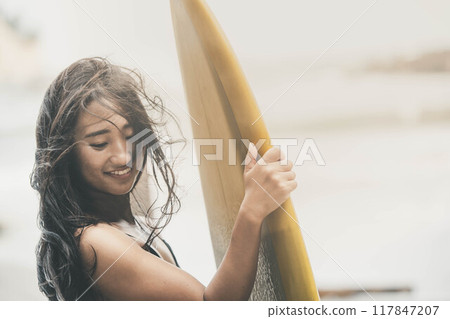 Young woman standing with surfboard in windy ocean 117847207