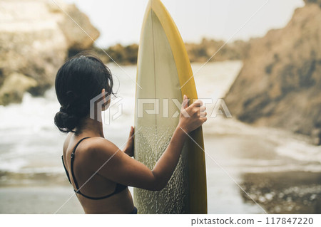 在風大的大海中拿著衝浪板站立的年輕女子 在風大的大海中拿著衝浪板站立的年輕女子 117847220