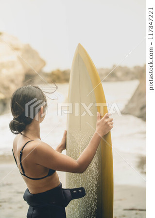 在風大的大海中拿著衝浪板站立的年輕女子 在風大的大海中拿著衝浪板站立的年輕女子 117847221