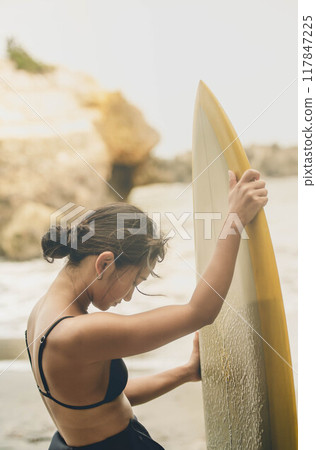 Young woman standing with surfboard in windy ocean 117847225